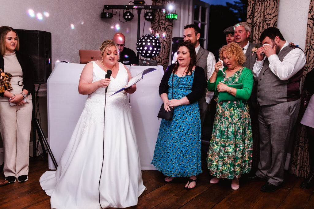 A bride in a white wedding dress reads from a paper into a microphone, surrounded by guests indoors, some listening attentively and others visibly emotional. DJ equipment is visible in the background.