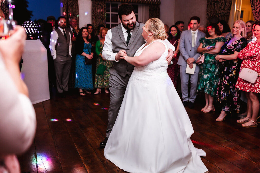 A bride and groom share their first dance at their wedding reception, smiling at each other, surrounded by guests who are watching and celebrating on a dance floor.