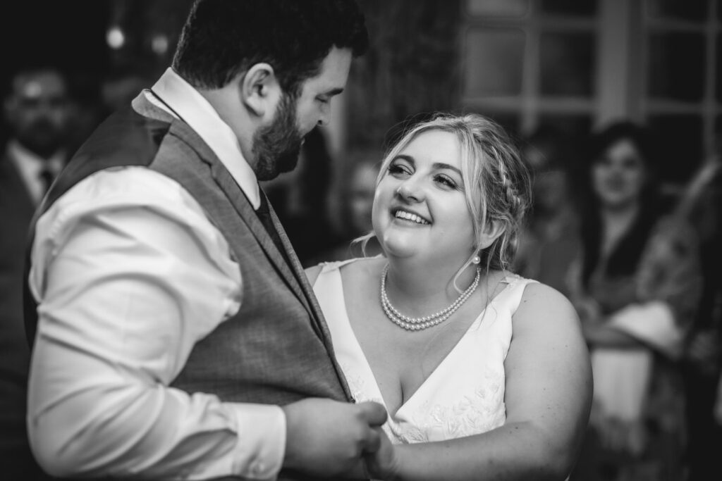 A bride and groom share a joyful moment while dancing together; the bride smiles warmly at the groom as they hold hands, with guests blurred in the background. The image is in black and white.