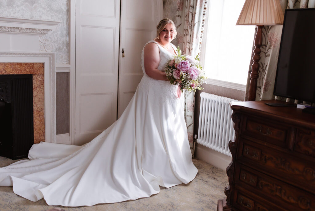 A bride in a white wedding dress with a long train stands by a window, holding a bouquet of flowers and smiling. The room has elegant decor, including a fireplace, a dresser, and a lamp.