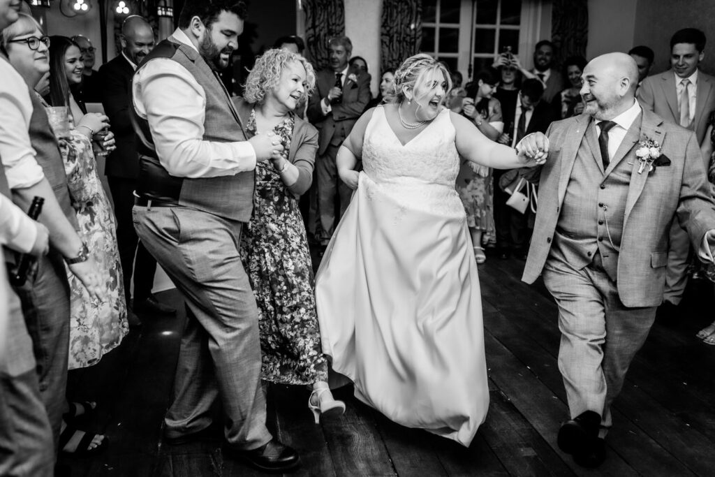 A joyful wedding scene shows two couples dancing together, smiling and holding hands, surrounded by guests in formal attire who watch and celebrate in a warmly lit indoor venue.