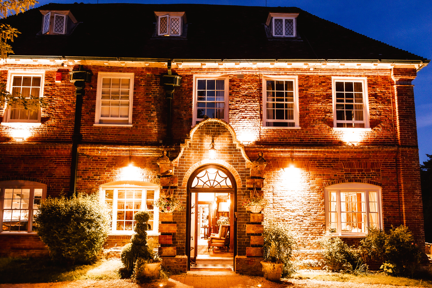 Two-story brick house lit warmly at night, with large windows, an arched doorway, and trimmed bushes framing the entrance. The interior light spills out, creating a welcoming atmosphere.