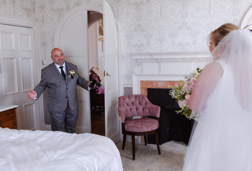 A man in a gray suit with a boutonniere smiles and opens his arms as he sees a bride in a white dress holding a bouquet, standing in a bright, elegant room with floral wallpaper and a pink chair.