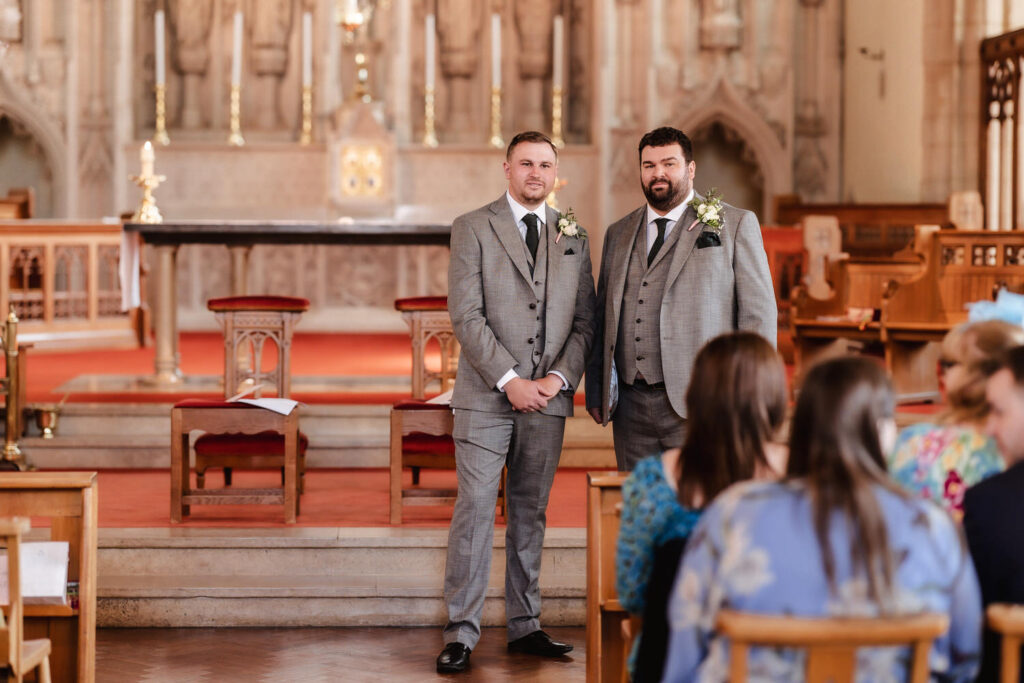 Two men in grey suits with boutonnieres stand at the front of a church, facing seated guests. The church interior features an ornate altar, wooden pews, and warm lighting.