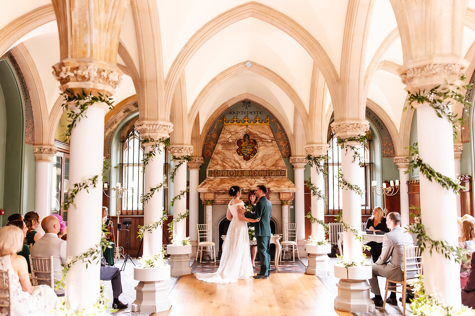 A bride and groom stand at the altar in a bright, elegant hall with arched ceilings and decorated columns, surrounded by seated guests during their wedding ceremony.