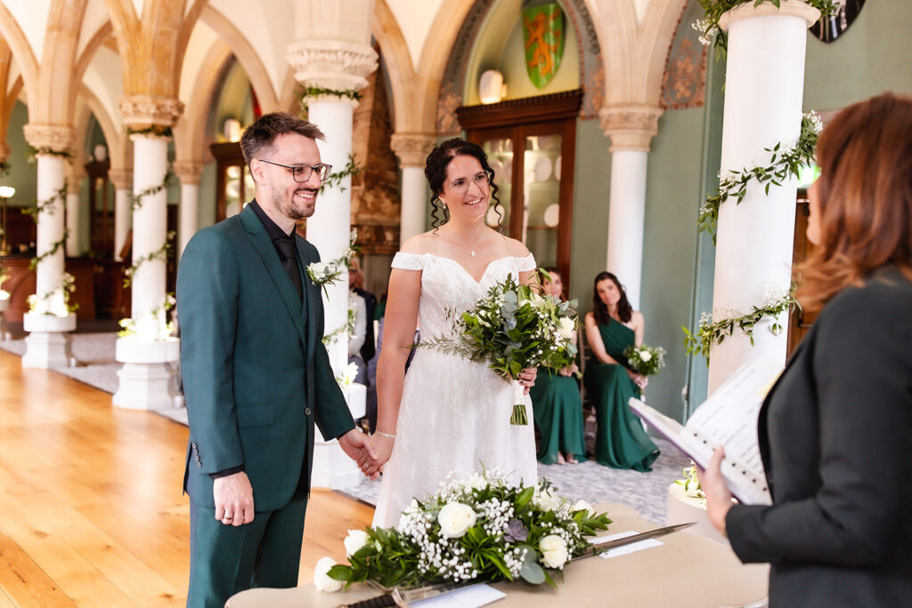 A bride and groom stand holding hands at a decorated table during a wedding ceremony in an elegant, columned hall, smiling at the officiant. Bridesmaids in green dresses sit in the background.