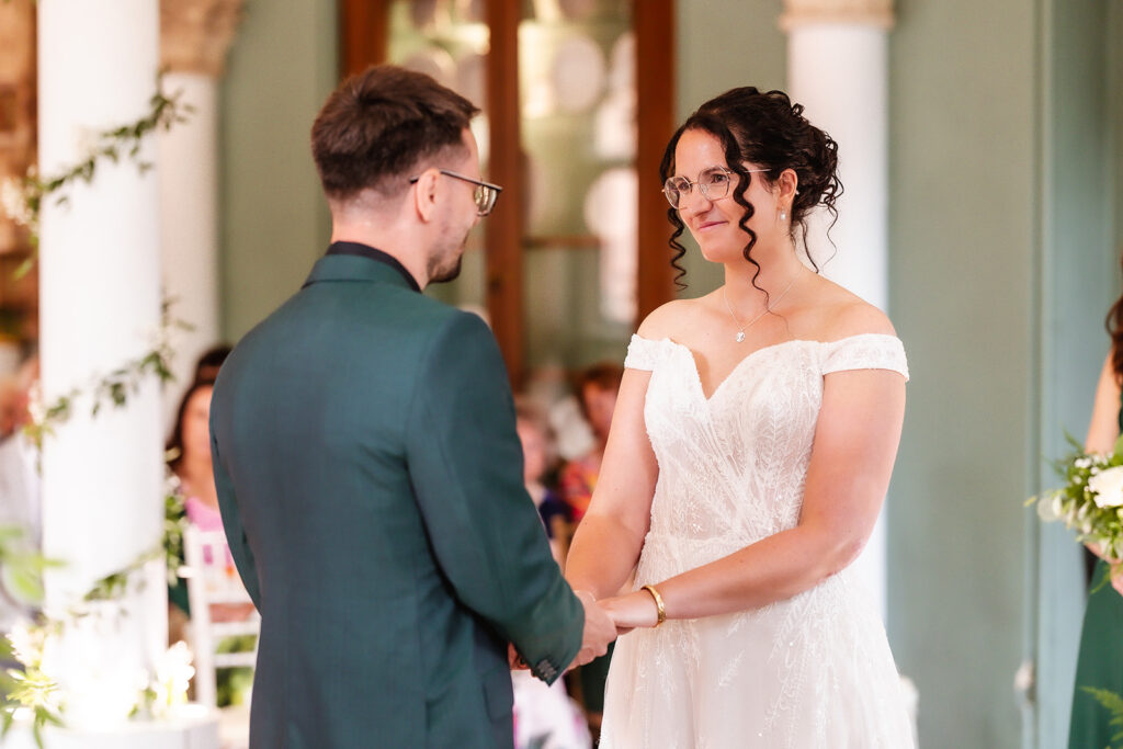 A bride in a white dress and glasses holds hands with a groom in a green suit during a wedding ceremony indoors, both looking emotional and smiling. White columns and blurred guests are visible in the background.