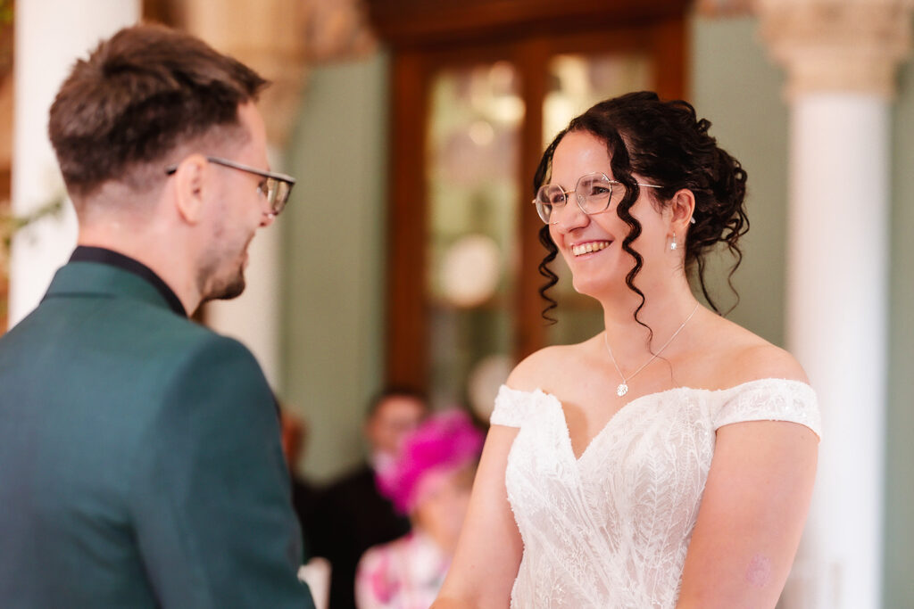 A bride in an off-shoulder white lace dress and glasses smiles at a man in a green suit jacket during a wedding ceremony in an elegant indoor setting.