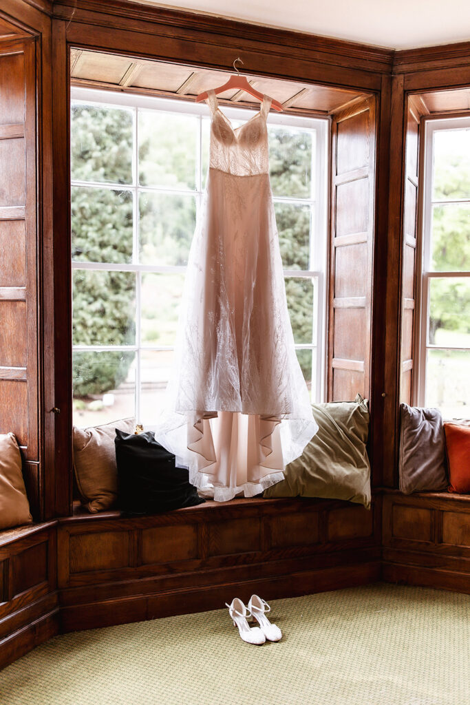 A white wedding dress hangs in a large window with wooden framing, above a cushioned window seat with pillows. White bridal shoes are placed on the floor below. Bright natural light fills the room.