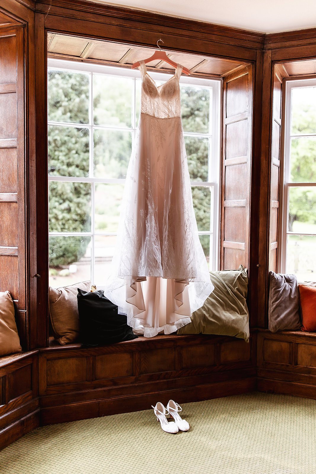 A white wedding dress hangs in a large window with wooden framing, above a cushioned window seat with pillows. White bridal shoes are placed on the floor below. Bright natural light fills the room.