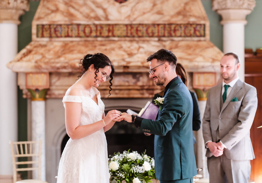 A bride and groom exchange rings during their wedding ceremony, standing in front of an ornate fireplace. The bride wears an off-shoulder white dress; the groom wears a dark green suit. A groomsman in a gray suit stands behind them.