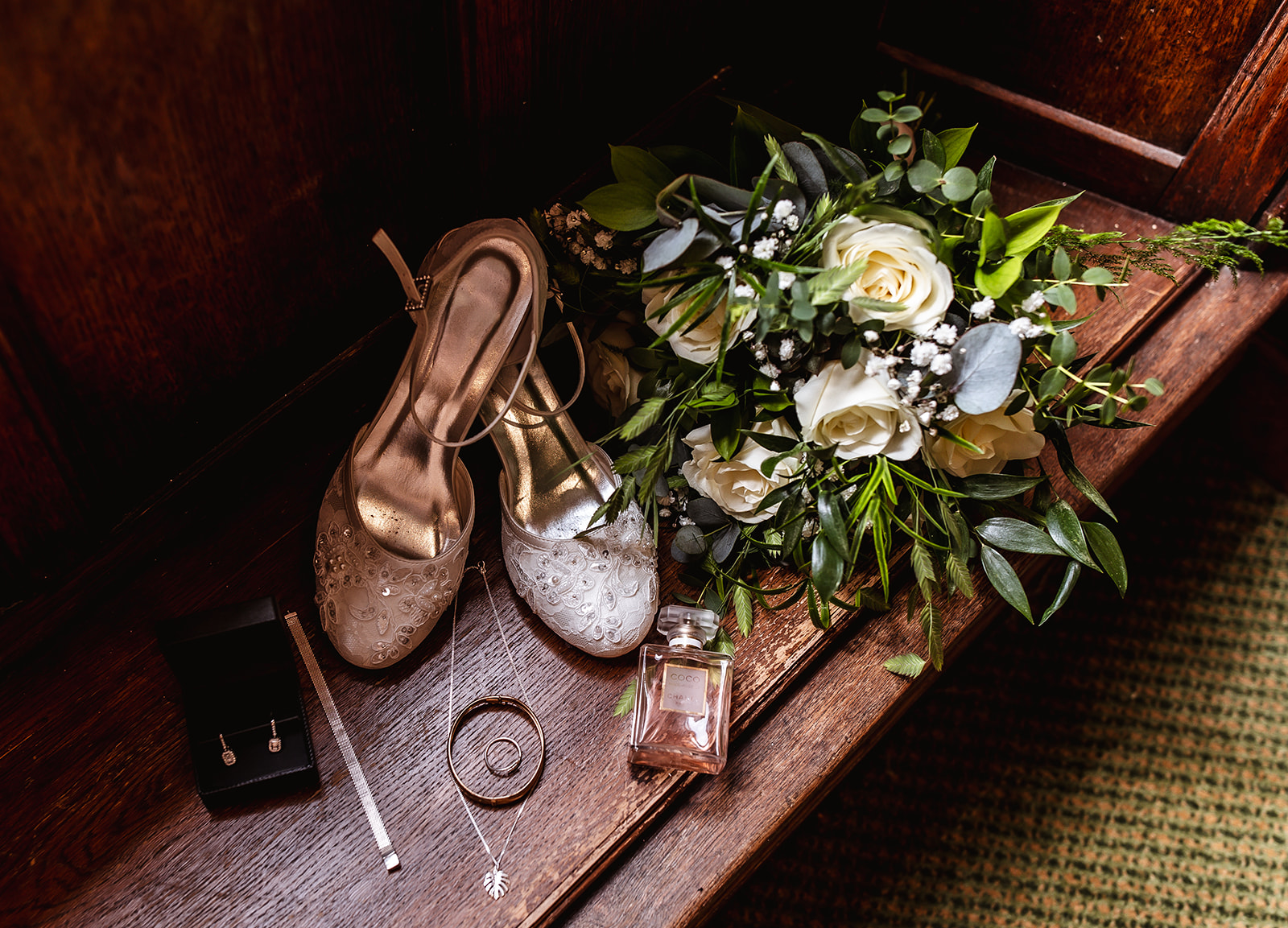Elegant bridal accessories arranged on a wooden bench: white floral bouquet, embellished high heels, perfume bottle, jewelry box with earrings, a bracelet, and a thin silver necklace.