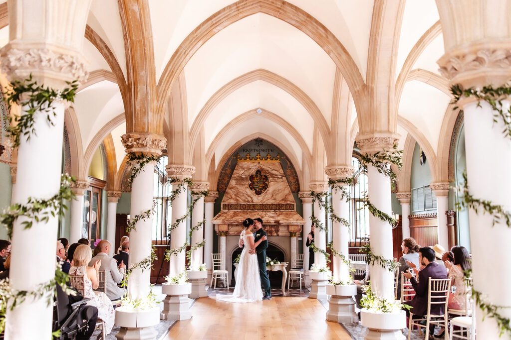 A bride and groom share their first kiss at the altar in a grand, arched hall decorated with greenery. Guests sit on white chairs, watching the couple under soaring columns and vaulted ceilings.