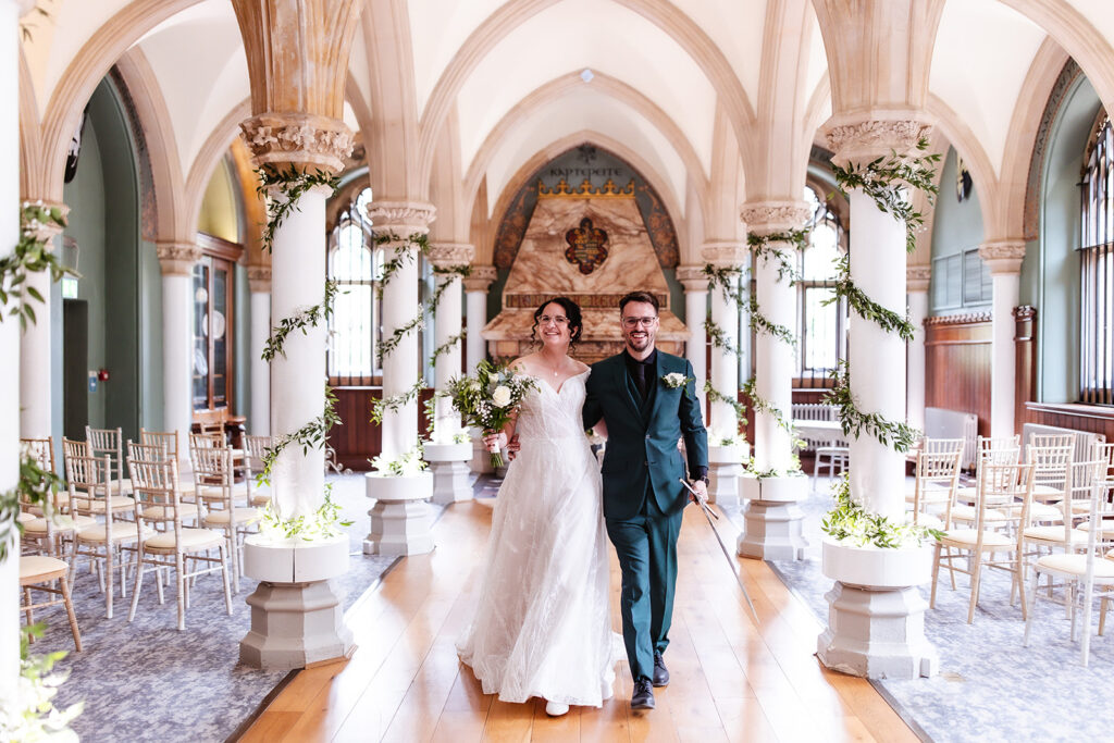 A bride and groom walk arm in arm down the aisle of an ornate hall decorated with greenery and white chairs, smiling happily after their wedding ceremony.