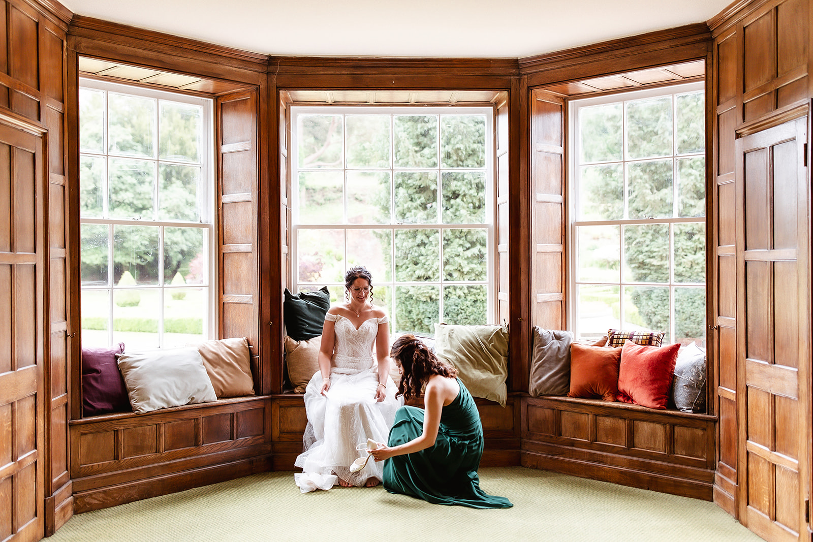 A bride sits on a window seat in a bay window with wooden paneling, smiling as another woman in a green dress kneels to adjust her shoe. Bright natural light fills the room, highlighting colorful cushions and a garden view.