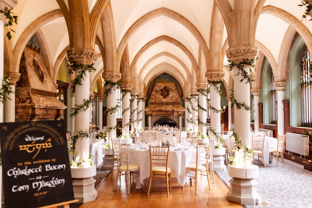 A grand hall with arched ceilings and columns decorated with greenery, set for a wedding reception with round tables covered in white linens and gold chairs; a sign welcomes guests to the wedding of Charlotte and Tom.