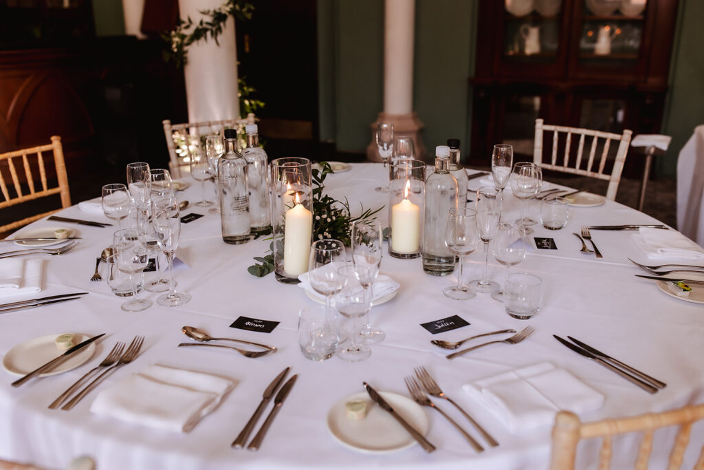 A round table set for a formal event with white tablecloth, glassware, plates, cutlery, water bottles, candles, and greenery centerpiece, surrounded by wooden chairs. Place cards are positioned at each setting.