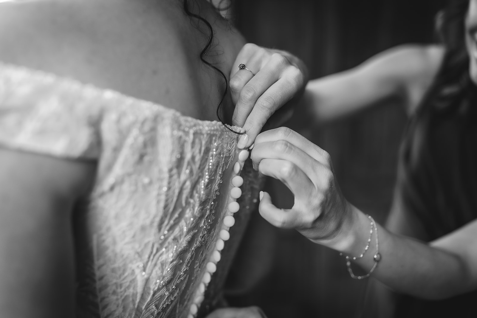 A close-up, black-and-white image shows someone fastening the buttons on the back of a bride’s lacy, off-the-shoulder wedding dress. Another person’s hands assist with the delicate task.