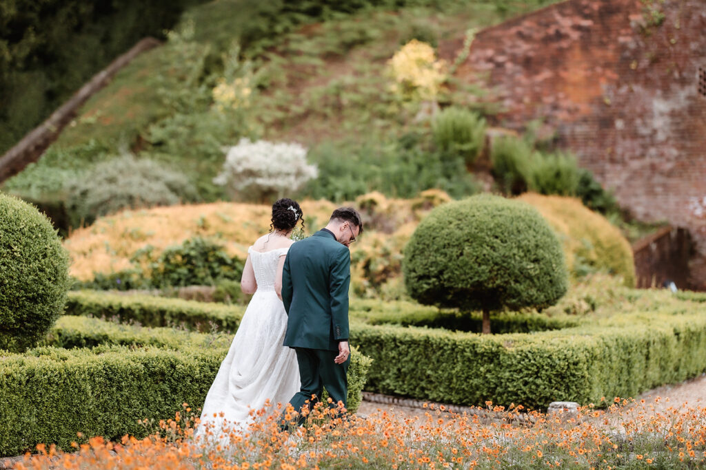 A bride and groom walk hand-in-hand through a manicured garden, surrounded by trimmed hedges, bright orange flowers, and lush greenery. The bride wears a white dress; the groom is in a dark suit.