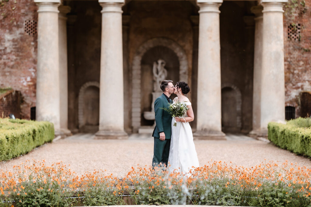 A couple in wedding attire kisses in front of tall stone columns and an arched structure, surrounded by blooming orange flowers and greenery. The bride holds a bouquet of flowers.
