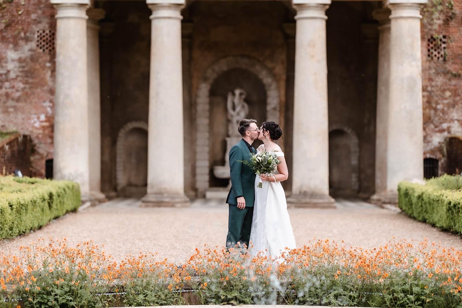 A newlywed couple kisses in front of tall stone columns, surrounded by blooming orange flowers and greenery. The bride holds a bouquet and wears a white dress; the groom is in a dark suit.