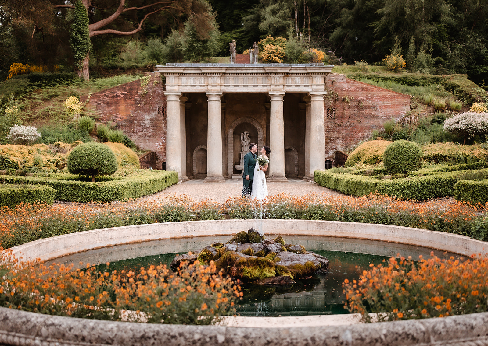 A bride and groom stand together in front of a classical stone structure, surrounded by vibrant orange flowers, manicured bushes, and a round fountain in a lush garden.