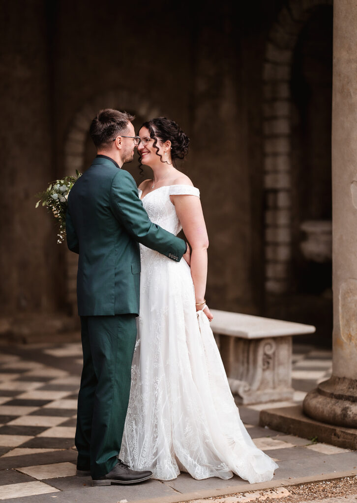 A bride and groom stand close together, smiling at each other. The bride wears an off-the-shoulder white gown and holds a bouquet. The groom wears a dark green suit. They are outdoors on a stone floor with columns and arches behind them.