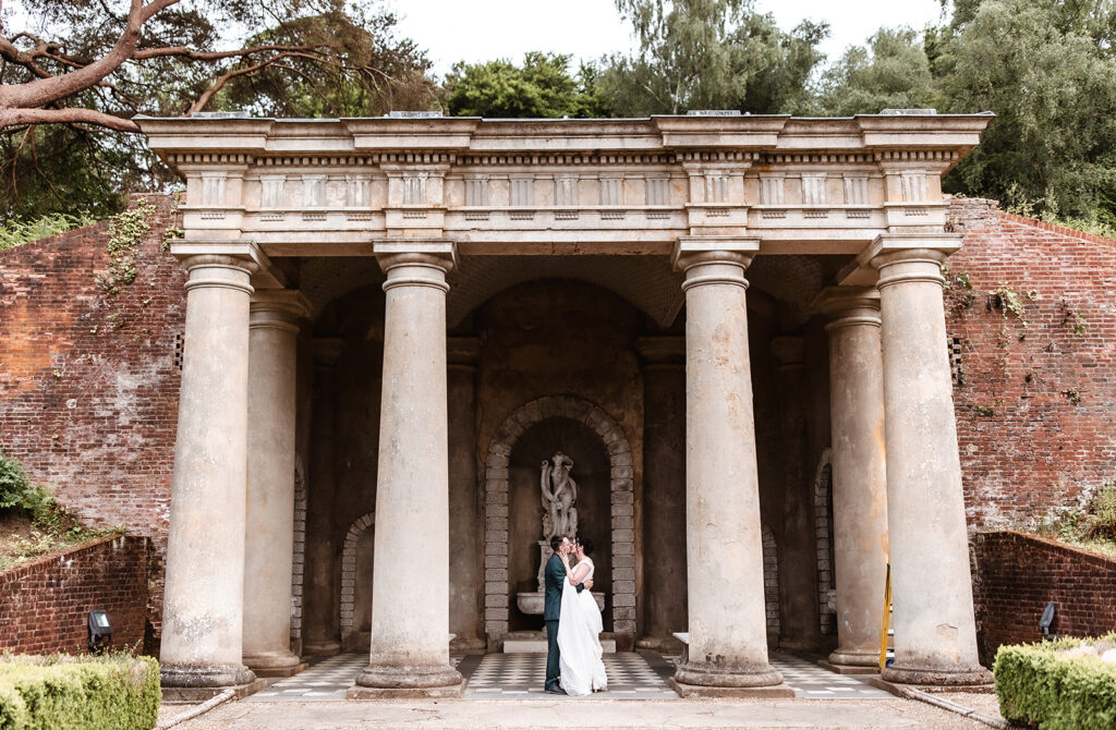 A bride and groom embrace in front of a grand, classical stone pavilion with tall columns and a brick wall, surrounded by greenery and an arched statue in the background.