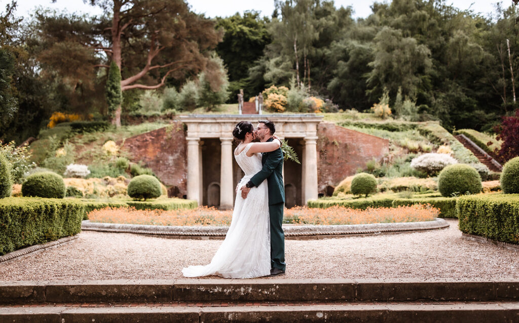 A bride and groom embrace and kiss on a gravel path in a formal garden, with lush greenery, manicured hedges, and a classical stone pavilion in the background.