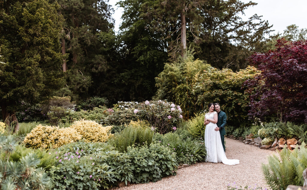 A newlywed couple stands embracing on a gravel path in a lush garden surrounded by vibrant green plants and blooming flowers, with tall trees in the background.