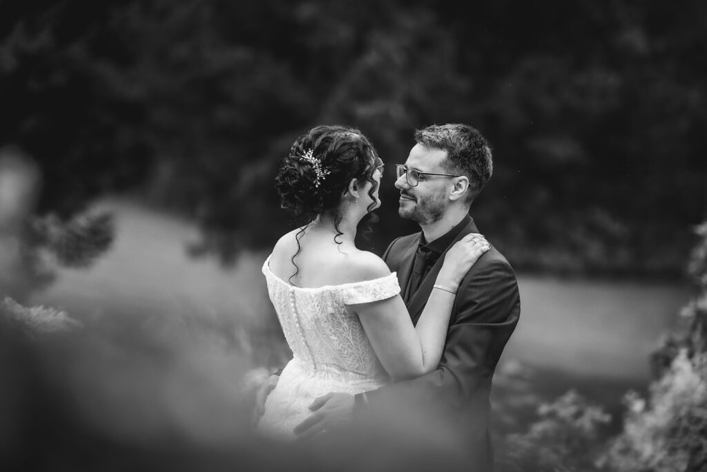 A black and white photo of a bride in an off-the-shoulder dress and a groom in a suit, smiling and embracing each other outdoors, surrounded by blurred foliage.