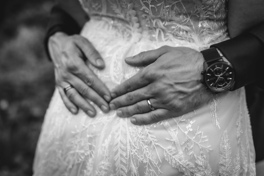 Close-up of two hands, both wearing wedding rings, gently resting on the back of a person in a lace wedding dress. One hand has a large wristwatch. The image is in black and white.