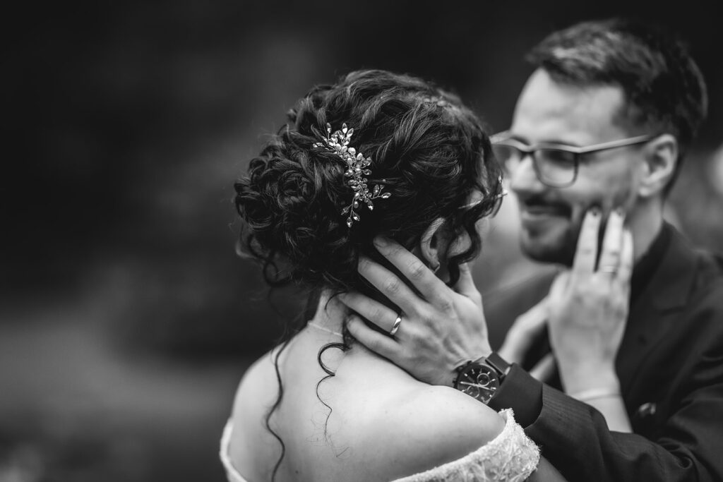A black and white photo of a bride and groom in an intimate moment. The bride has curly hair with a decorative hairpiece and gently holds the groom’s face, while the groom smiles and touches her hands.