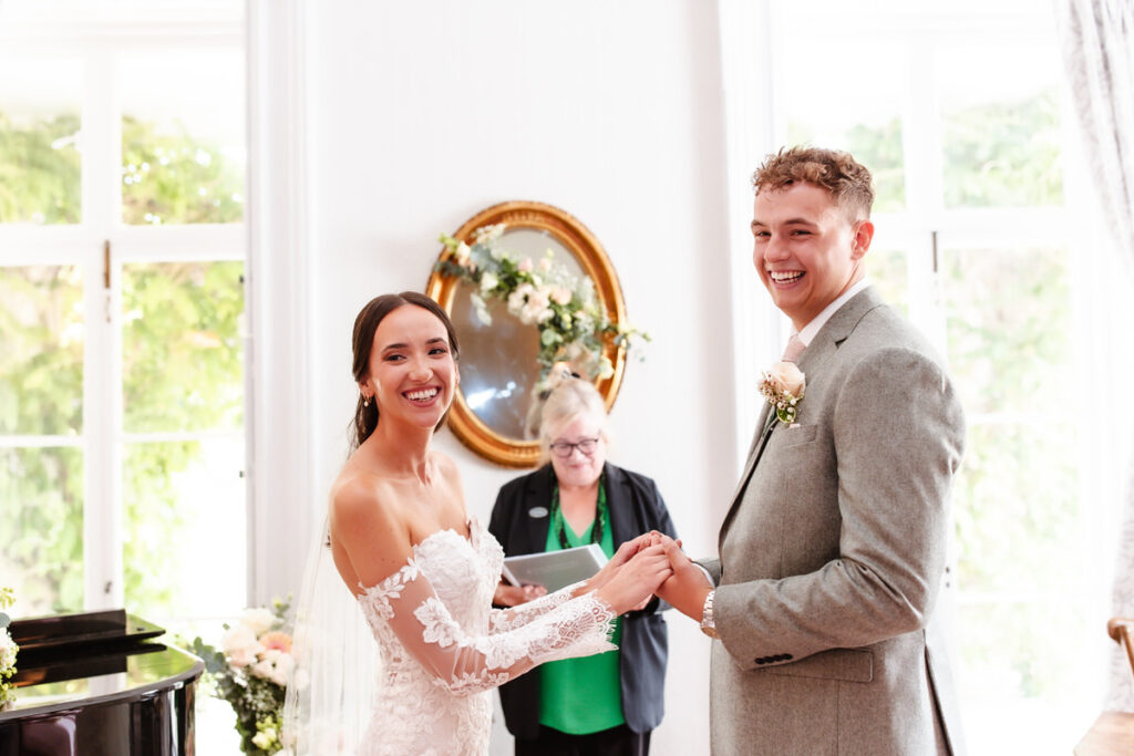 A bride and groom smile joyfully while holding hands during their wedding ceremony, with an officiant standing behind them in a bright, elegant room decorated with flowers and a gold-framed mirror.
