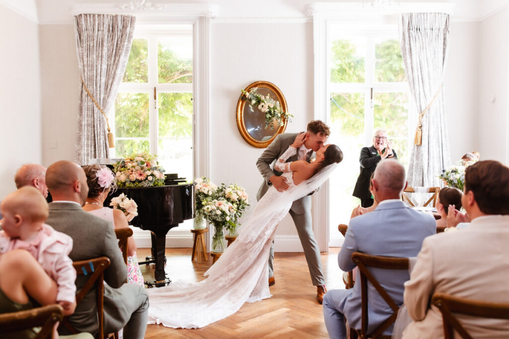 A bride and groom kiss at the altar in a bright room as guests watch and applaud. The bride wears a long white dress; flowers and a piano decorate the space. A photographer captures the moment near large windows with curtains.