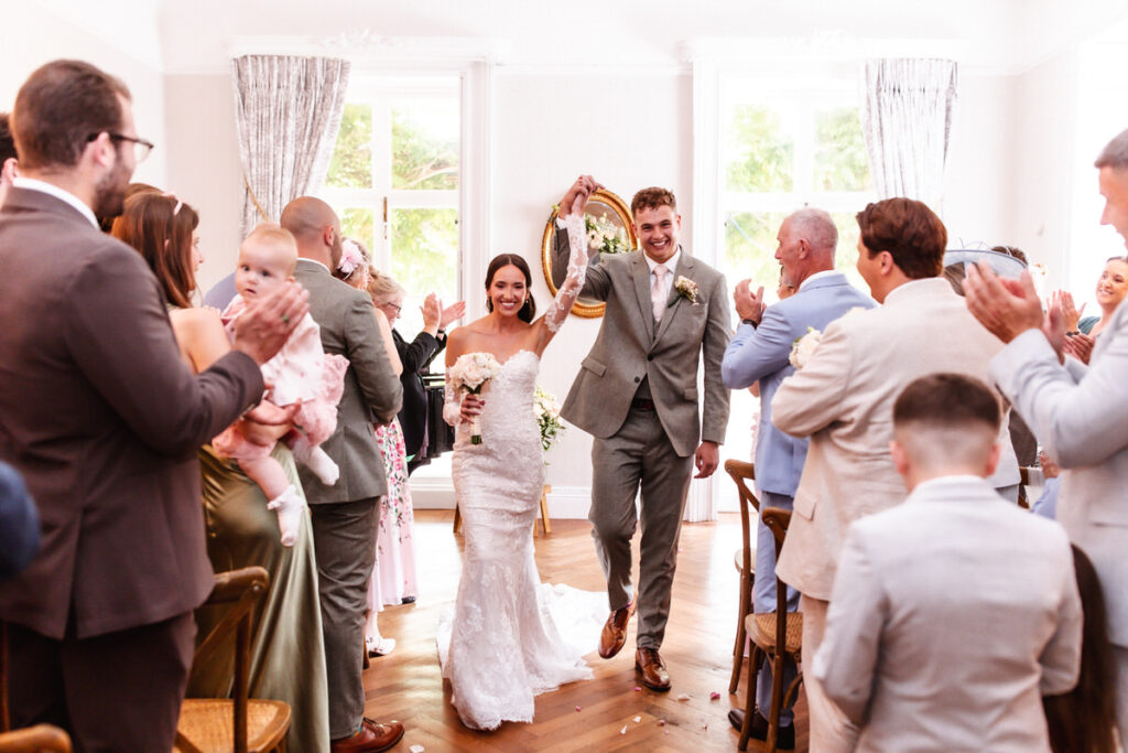A smiling bride and groom walk down the aisle together, holding hands, while guests on both sides clap and celebrate their wedding. The room is bright with natural light from large windows behind them.