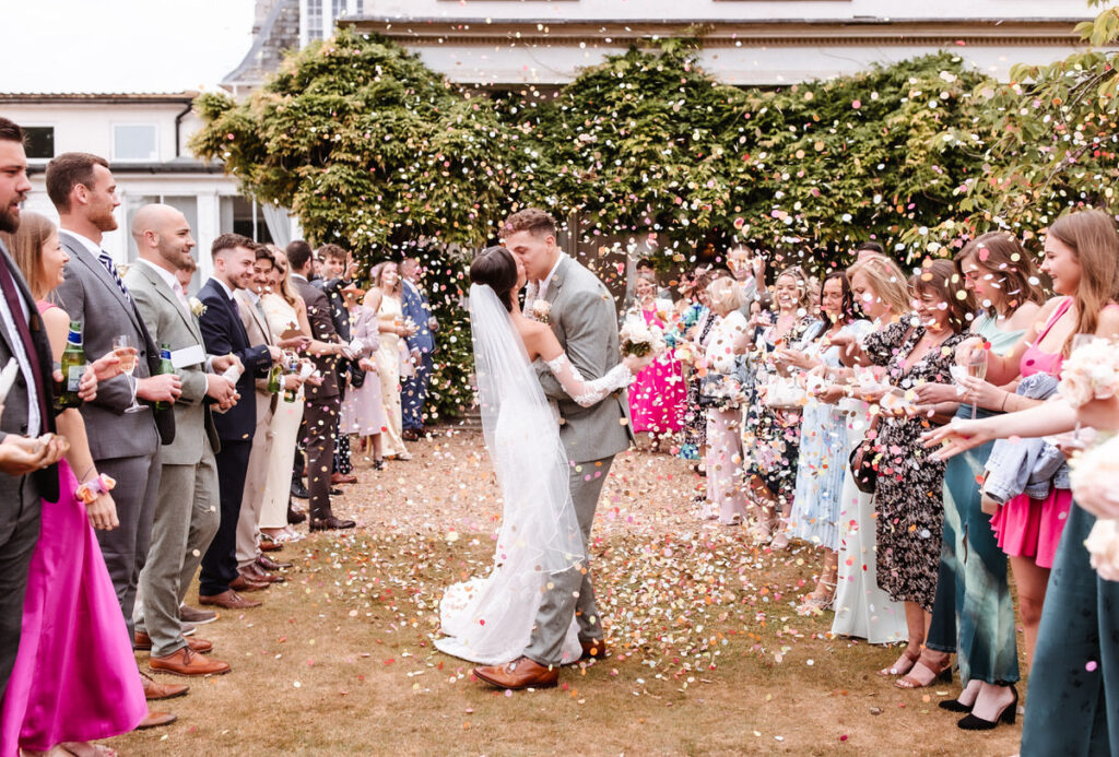 A bride and groom kiss outdoors as wedding guests on both sides throw confetti. The couple stands in the center, surrounded by smiling, well-dressed guests under a green leafy backdrop.