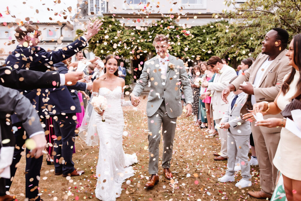 A newlywed couple walks hand-in-hand outdoors as guests throw flower petals over them, celebrating their wedding. Both are smiling and surrounded by cheering friends and family.