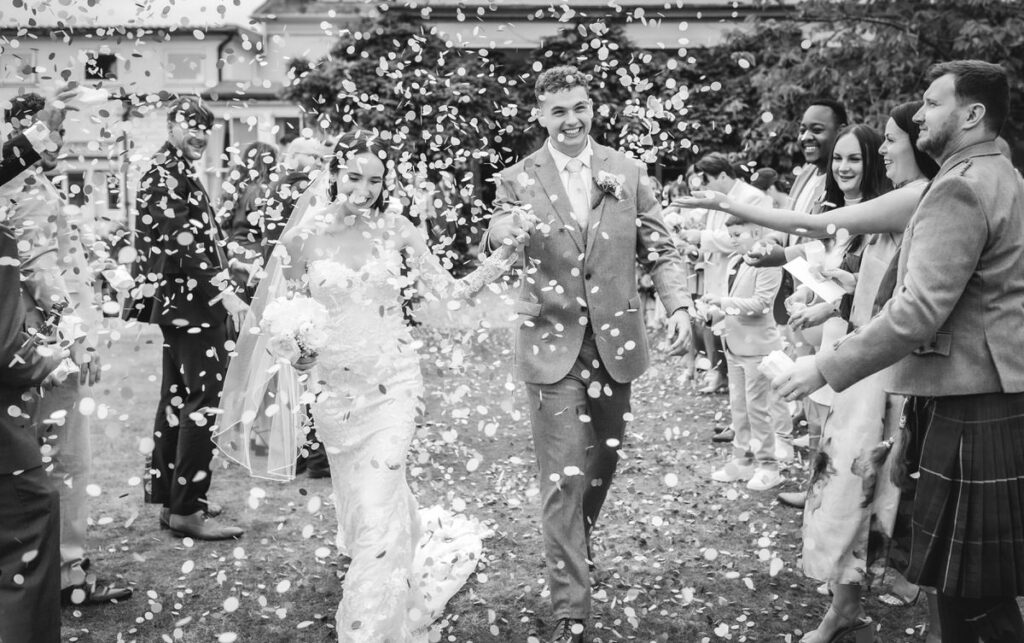 A bride and groom smile and hold hands while guests throw flower petals over them in celebration. The scene is joyful, lively, and takes place outdoors, with the wedding party surrounding the couple. The image is in black and white.