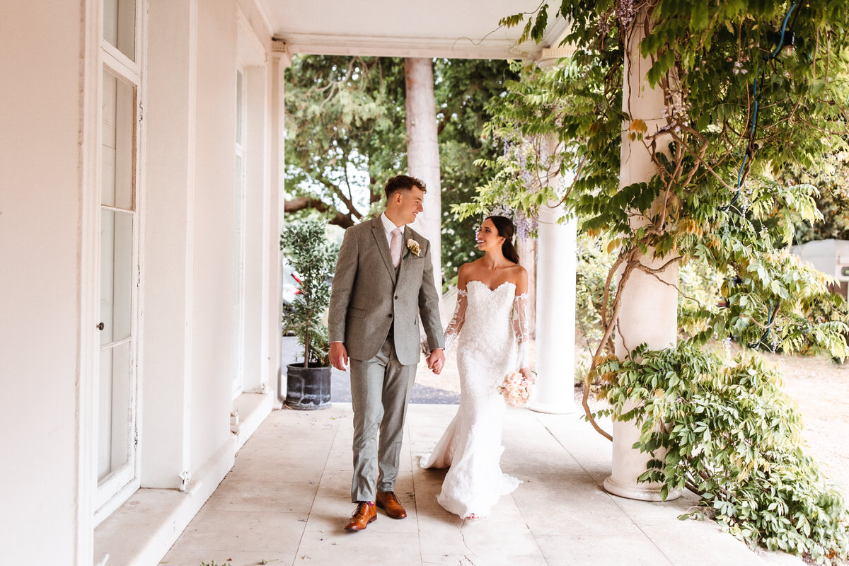 A bride in a white, strapless gown and a groom in a light gray suit walk hand-in-hand on a columned veranda, surrounded by greenery, smiling at each other.