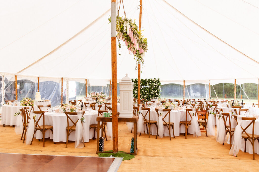 Elegant wedding reception under a large white tent with round tables covered in white linens, wooden chairs, floral centerpieces, and hanging greenery. Warm natural light fills the space.