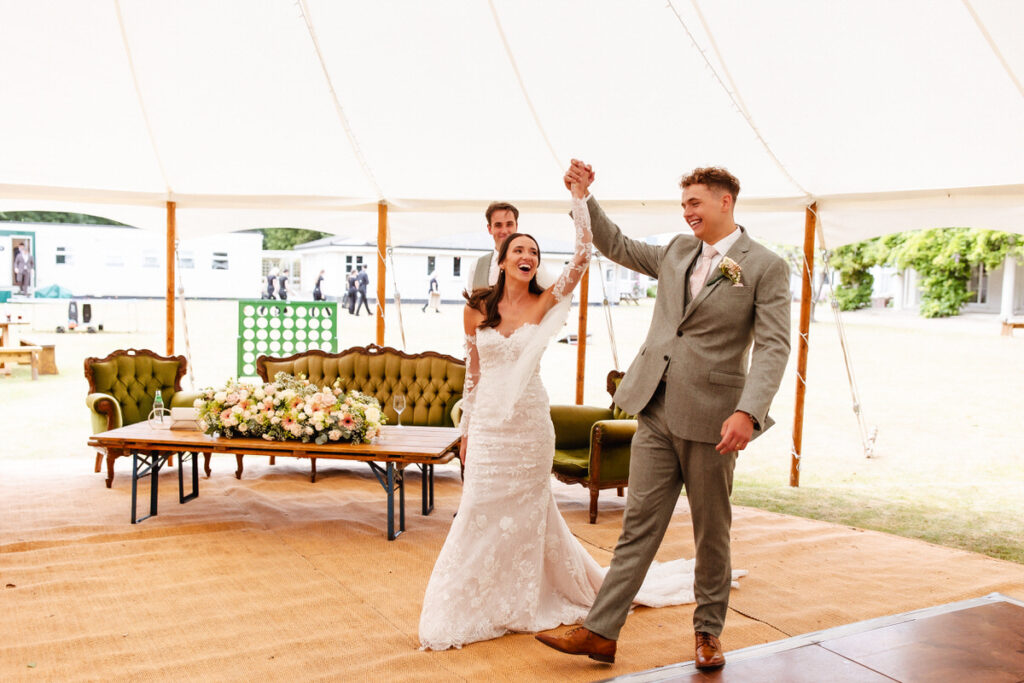 A bride and groom celebrate under a white tent, holding hands and smiling joyfully. The bride wears a white dress, the groom a light suit. Floral arrangements and vintage couches decorate the background.