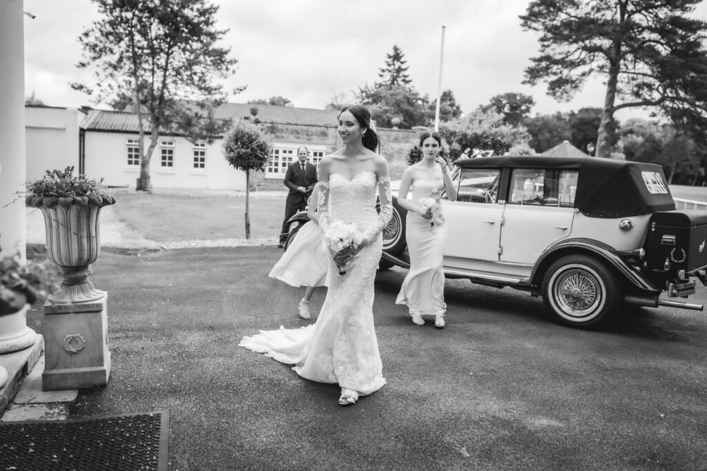 A bride in a long, lace wedding dress walks outside next to a vintage car, holding a bouquet, with two bridesmaids in light-colored dresses following her. The scene is outdoors near a building and trees.