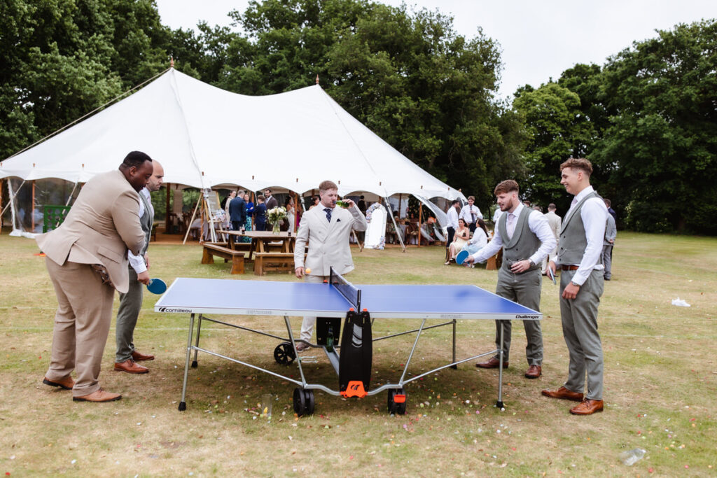 Five men dressed in suits and dress shirts play table tennis on a lawn in front of a large white event tent, with other guests and trees visible in the background.