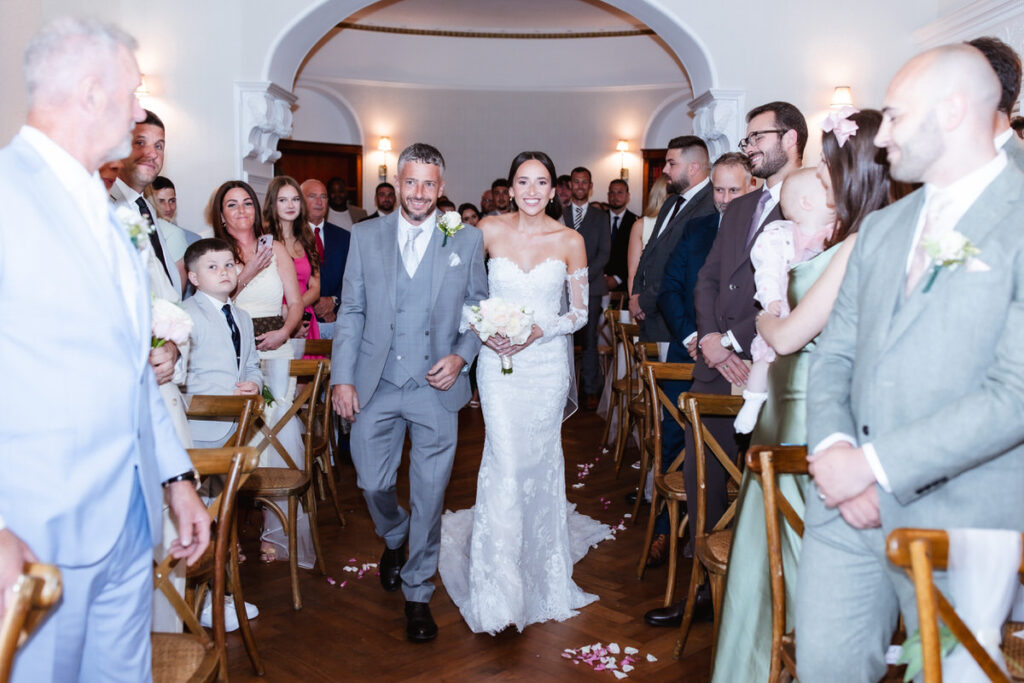 A bride in a white gown walks down the aisle with a man in a grey suit, smiling, as guests seated on either side watch and smile in a well-lit indoor venue.