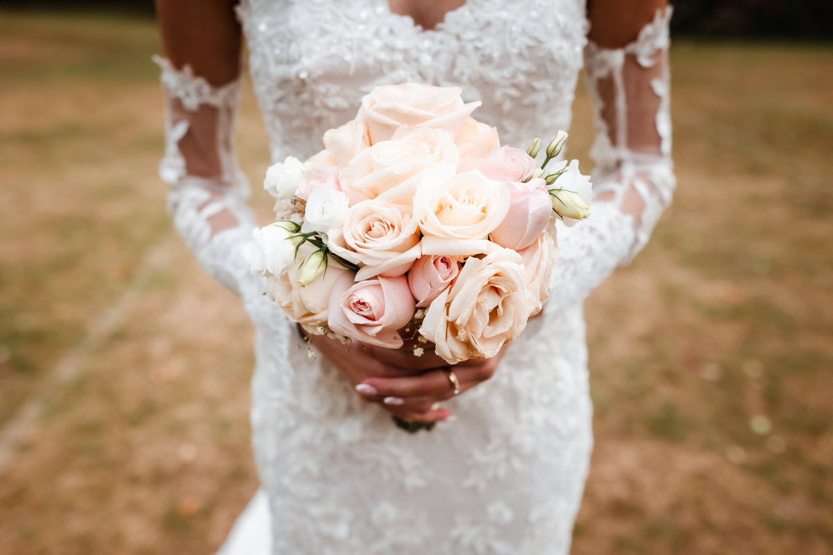 A bride in a white lace wedding dress holds a bouquet of pale pink and white roses, standing outdoors on a grassy field.