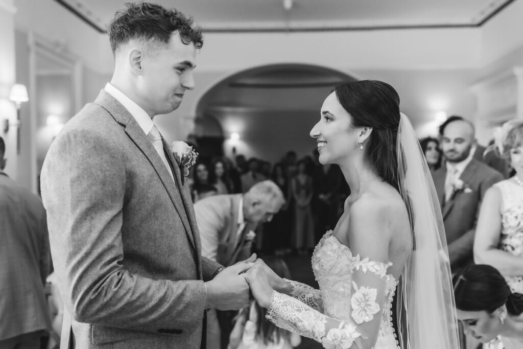 A bride and groom stand facing each other, holding hands and smiling during their wedding ceremony, with guests seated in the background. The image is in black and white.