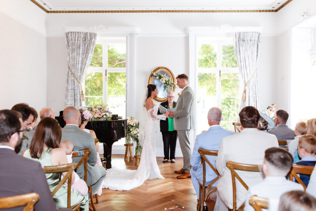 A bride and groom stand facing each other, holding hands during their wedding ceremony in a bright room with large windows, floral decor, and guests seated watching them.