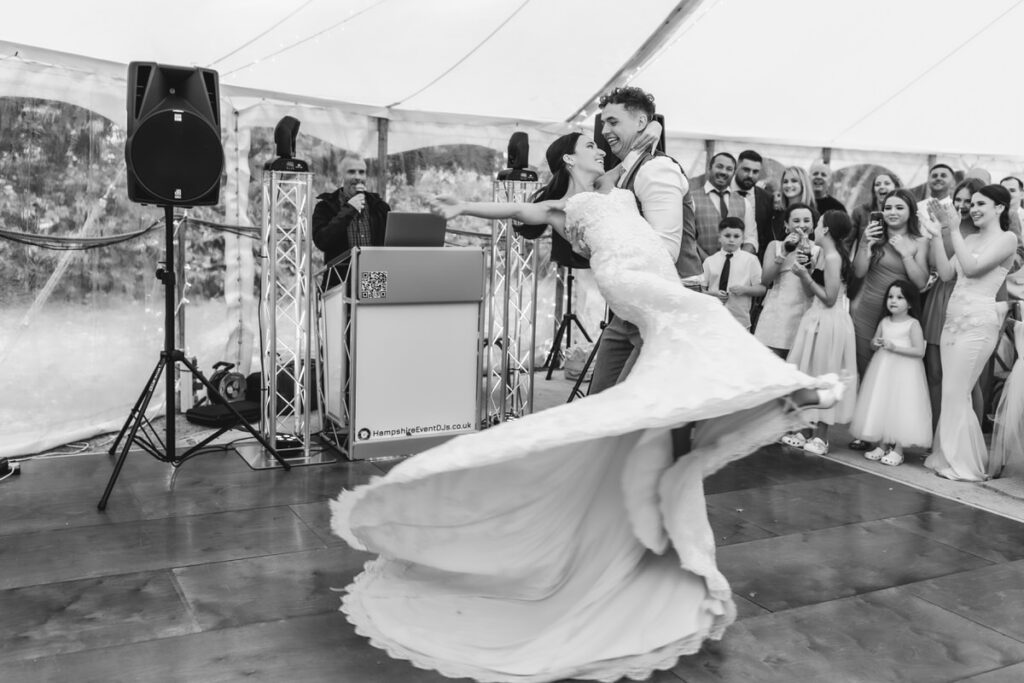 A groom twirls his bride on the dance floor as guests watch and smile, capturing a joyful moment at a wedding reception under a tent.