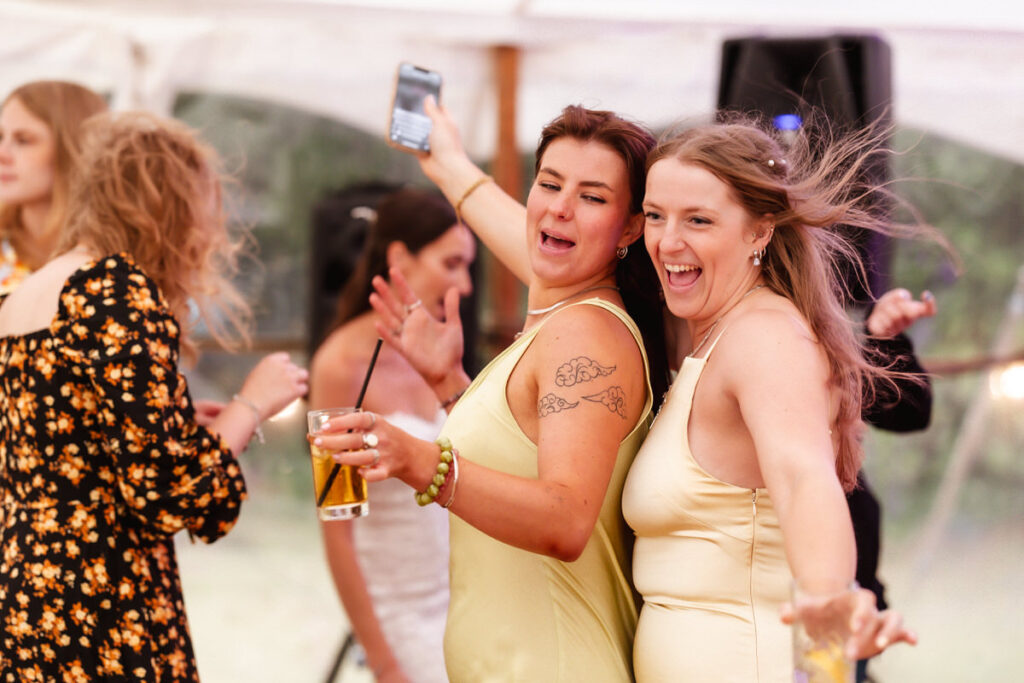 Two women in light yellow dresses smile and dance with their arms outstretched at a lively indoor event, while others dance in the background. One woman holds a drink, and both appear to be having fun.